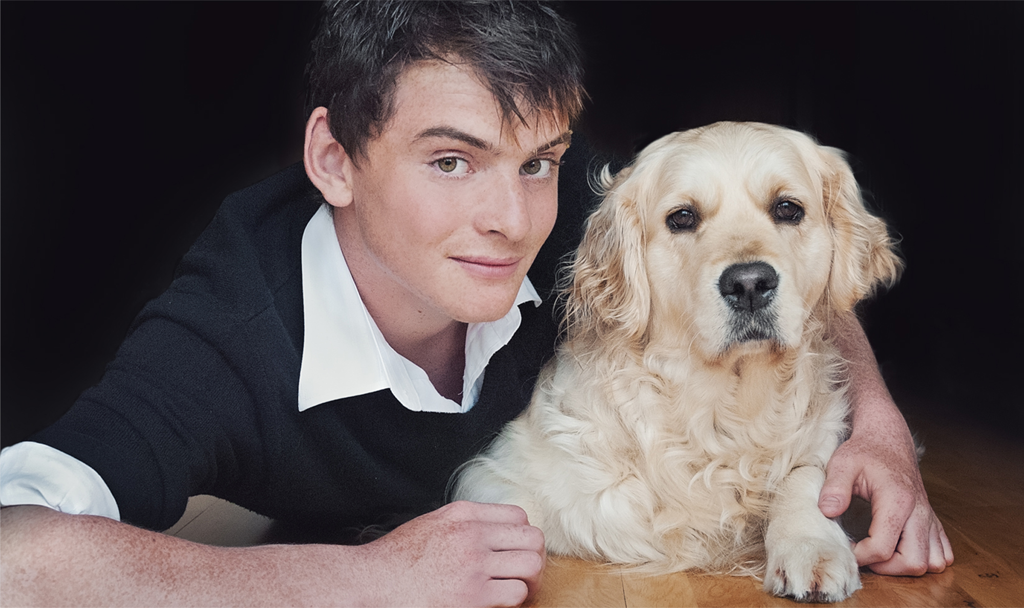 Family photos with dog_013 A young man in a black sweater and white shirt smiles while lying next to his golden retriever, capturing the essence of cherished family photos with a dog. Both have their heads close, with the canine looking directly at the camera. The dark background beautifully highlights their expressions.