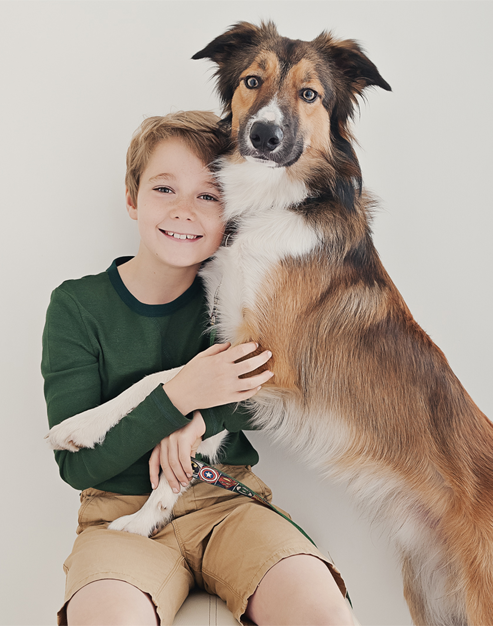 Family photos with dog_016 A smiling boy in a green shirt and khaki shorts sits with his brown-and-white dog, capturing the essence of family and dog photography. The dog's paws rest on the boy's lap as they both look happily at the camera against a plain background.