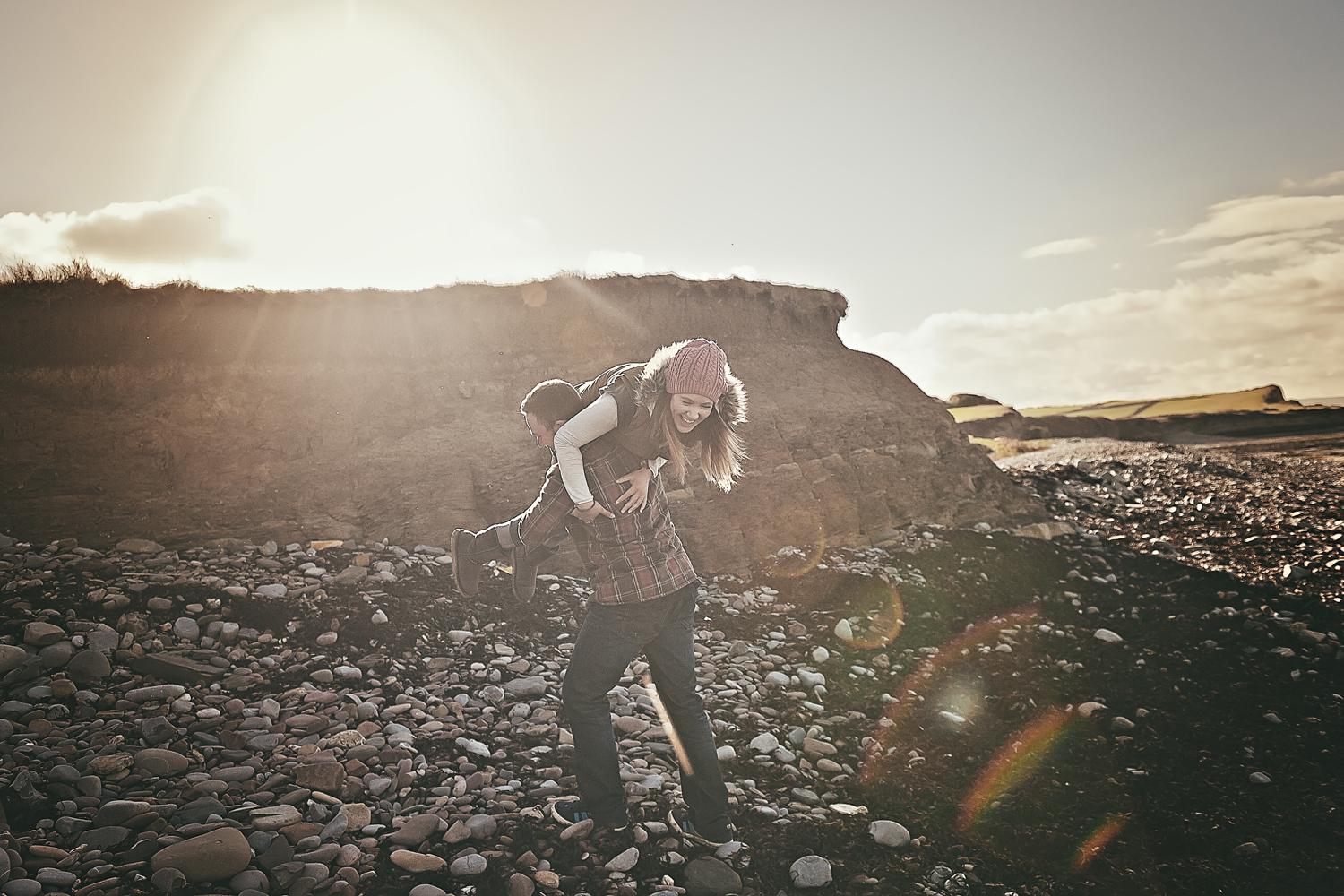 Playful, posed photograph of a woman lifted by her partner as the sun sets over Kilve Beach, Somerset.